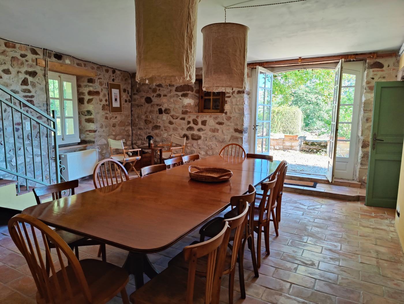 La salle à manger : grande table en bois, murs en pierre et porte-fenêtre ouverte sur le jardin