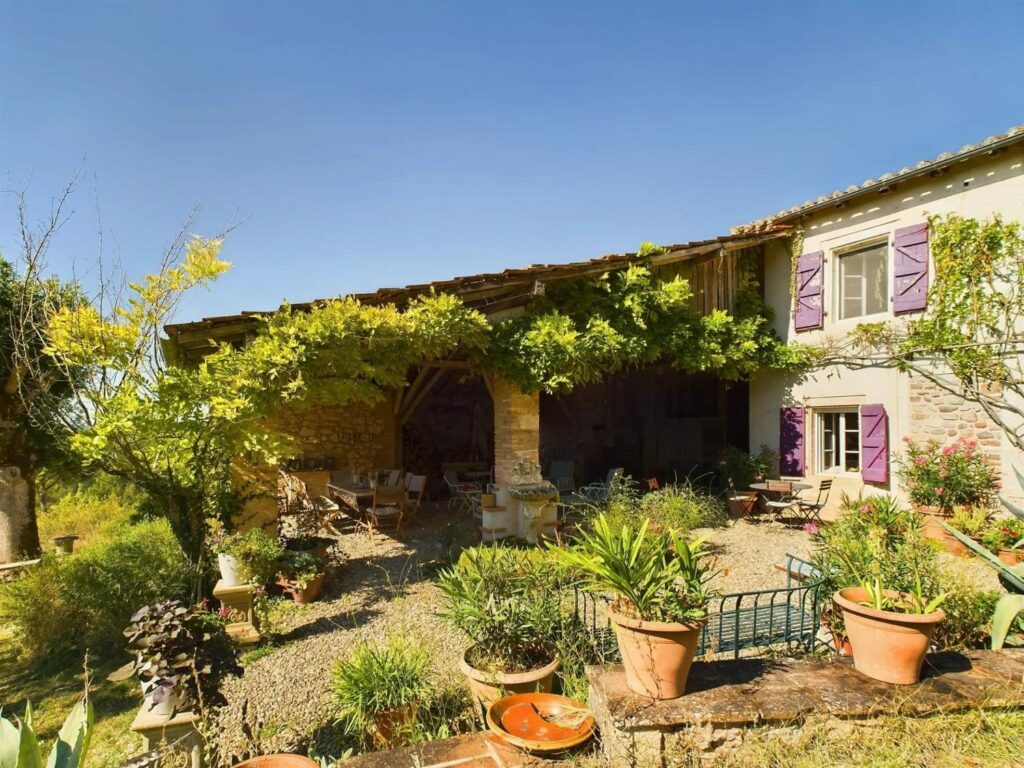 La façade et le préau couverts de glycine, avec les volets violets et des pots en terre cuite