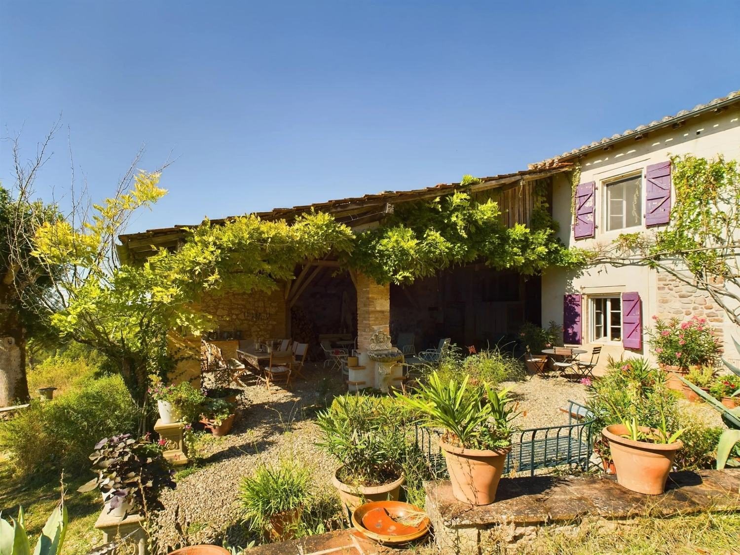 La façade et le préau couverts de glycine, avec les volets violets et des pots en terre cuite