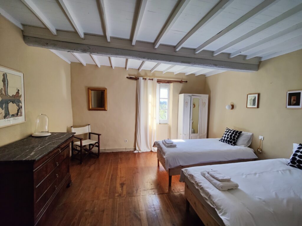 Grande chambre aux murs sable et poutres blanches, avec lits simples, commode ancienne et parquet